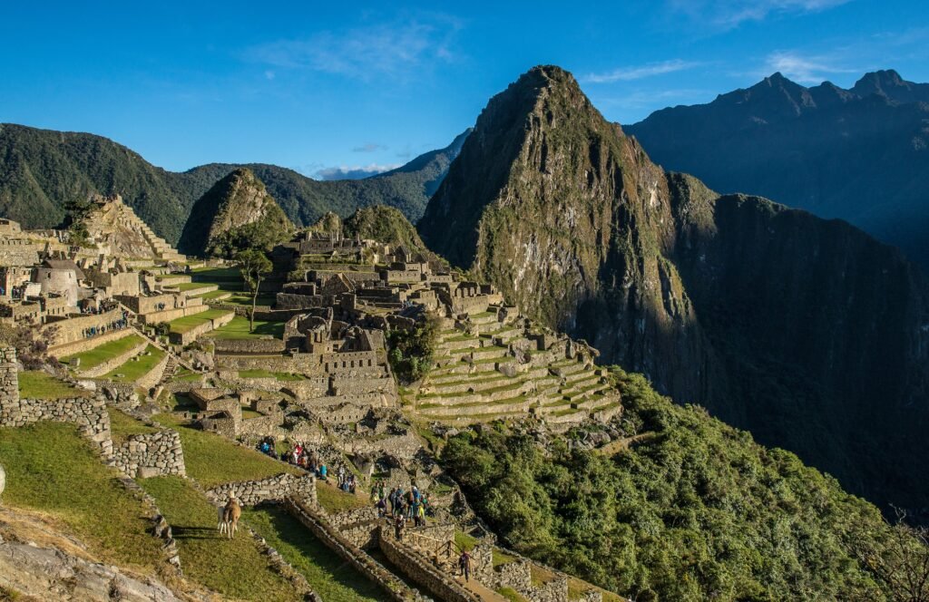 Ruínas de Machu Picchu com terraços agrícolas e montanhas imponentes ao fundo, sob um céu azul claro.