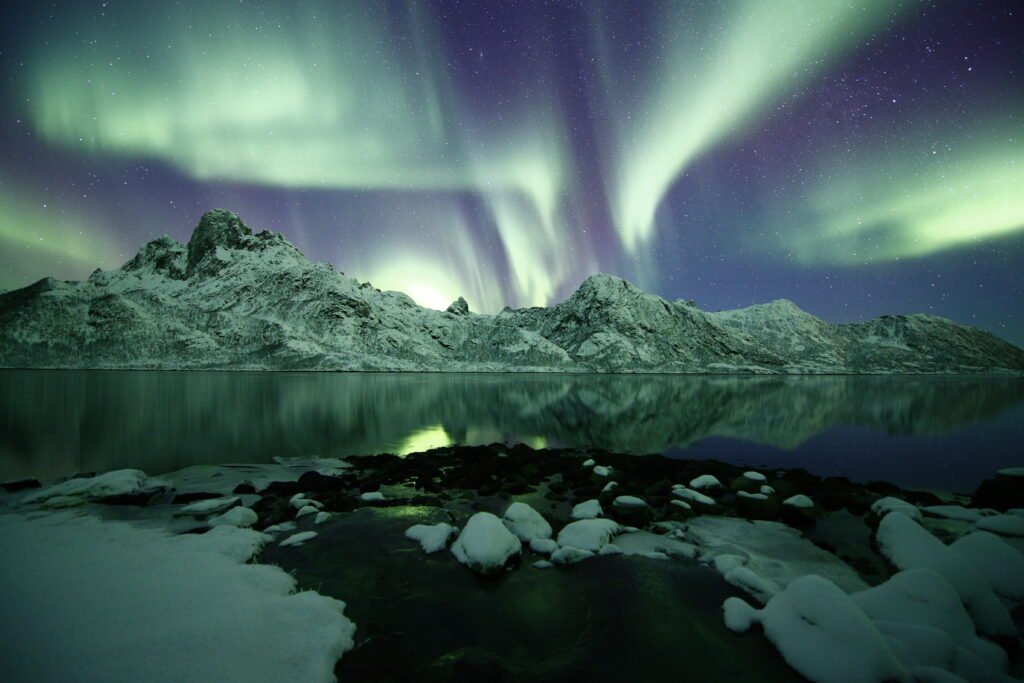 Auroras boreais iluminando o céu sobre montanhas nevadas e reflexos na água, representando fenômenos astronômicos em planetas.