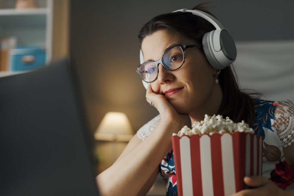 Mulher assistindo a um filme clássico em um laptop, com fones de ouvido e um balde de pipoca.